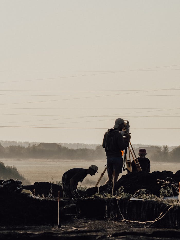 Group of surveyors using a theodolite at a construction site during a foggy morning with silhouettes visible.